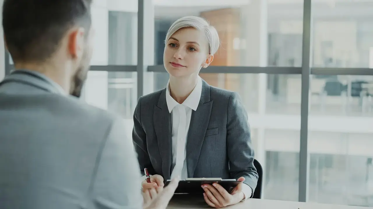 A job candidate speaking with an interviewer who is holding a clipboard in a bright office