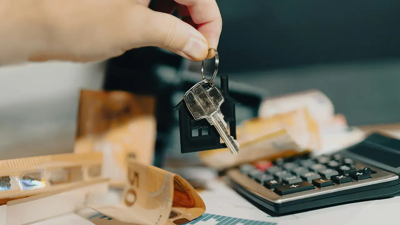 A hand holding a house-shaped keychain above a calculator and financial papers
